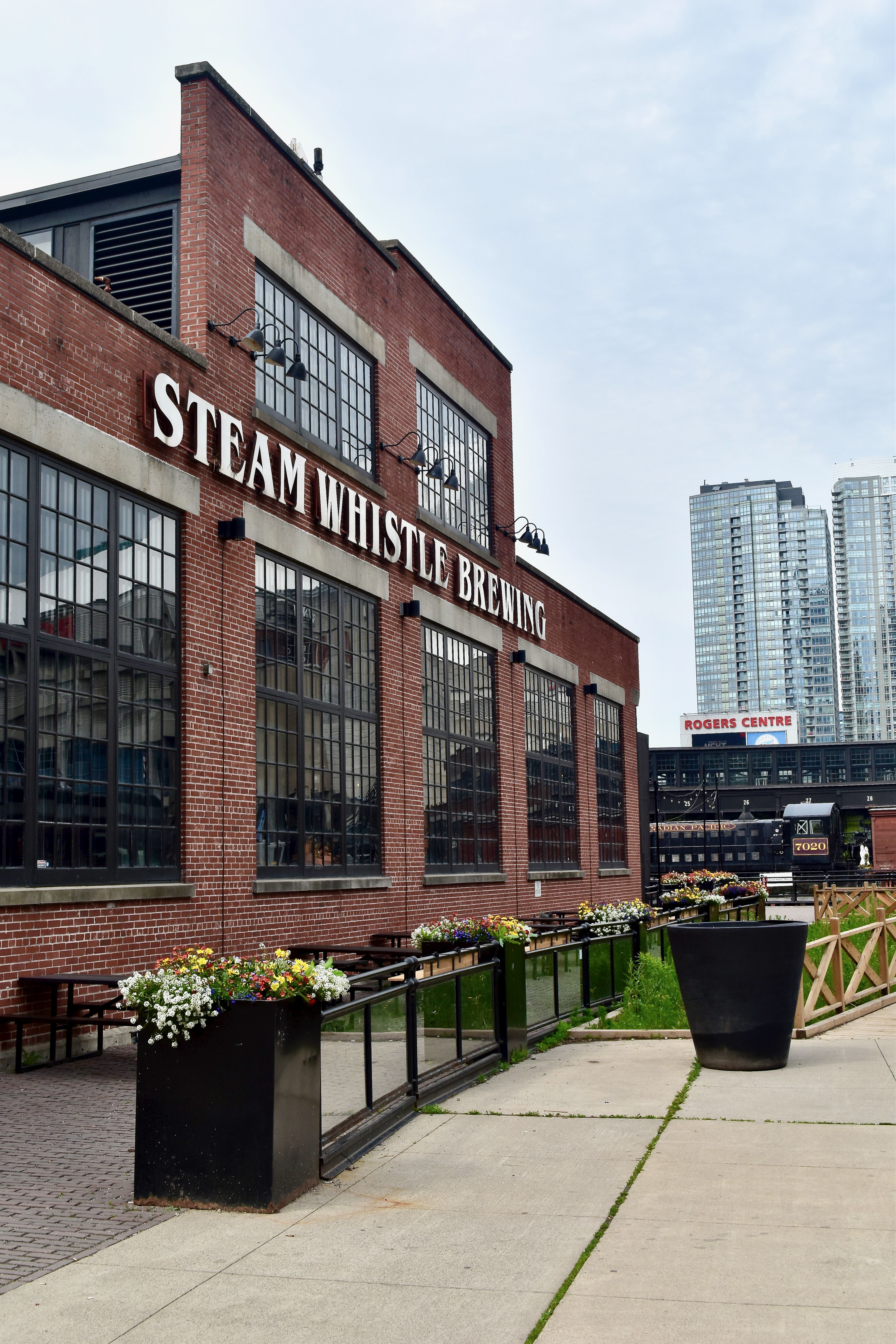 Large brick building with a series of large windows and old fashioned lights has a sign that reads, "Steam Whistle Brewing."