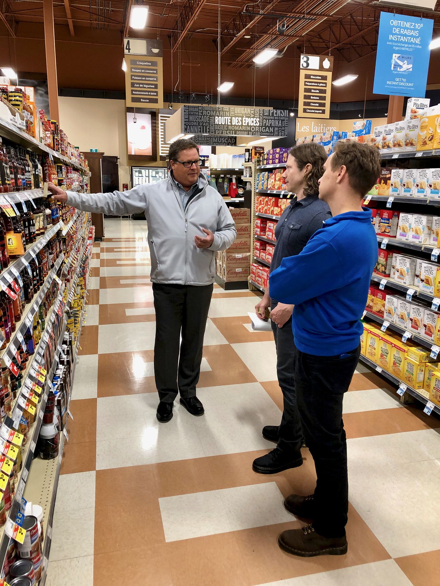Three men stand in the aisle of a grocery store looking at a large shelf of hot sauce options.