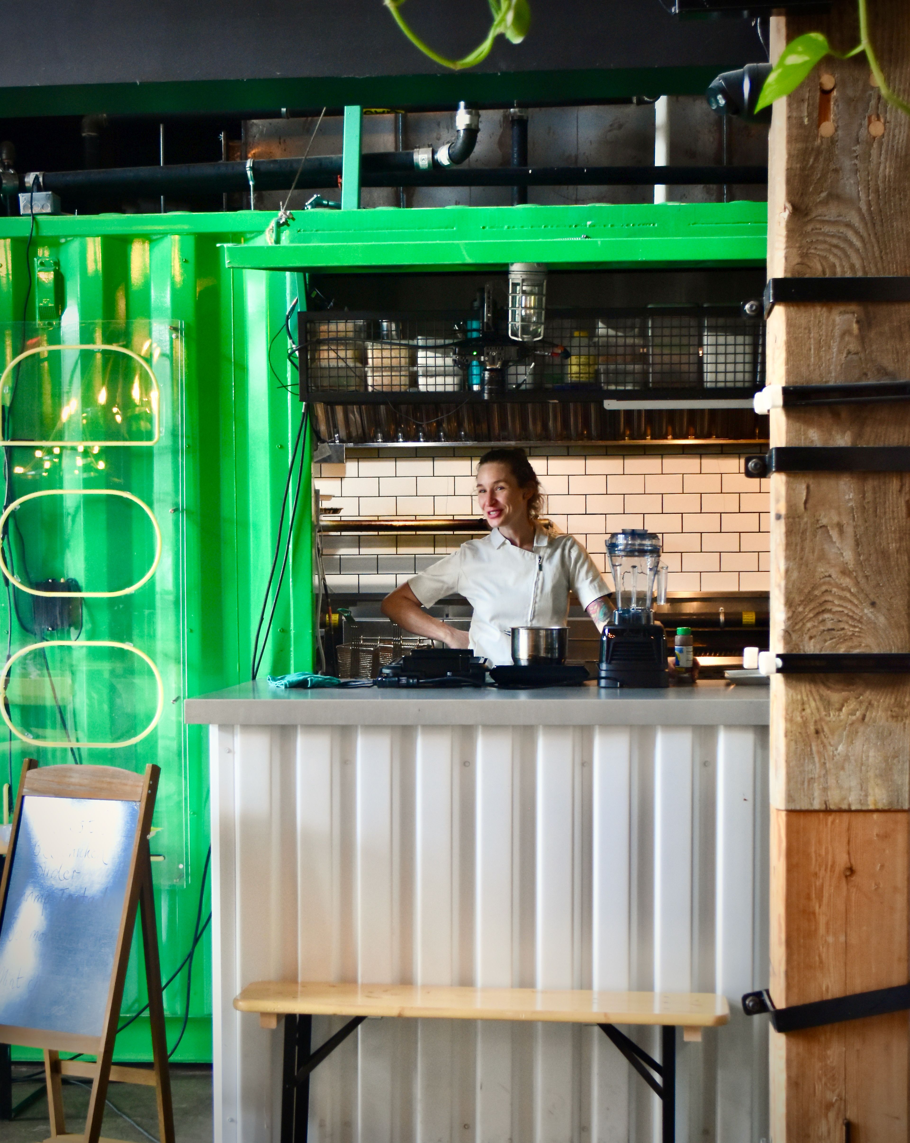 Woman stands behind a counter with a kitchen behind her.
