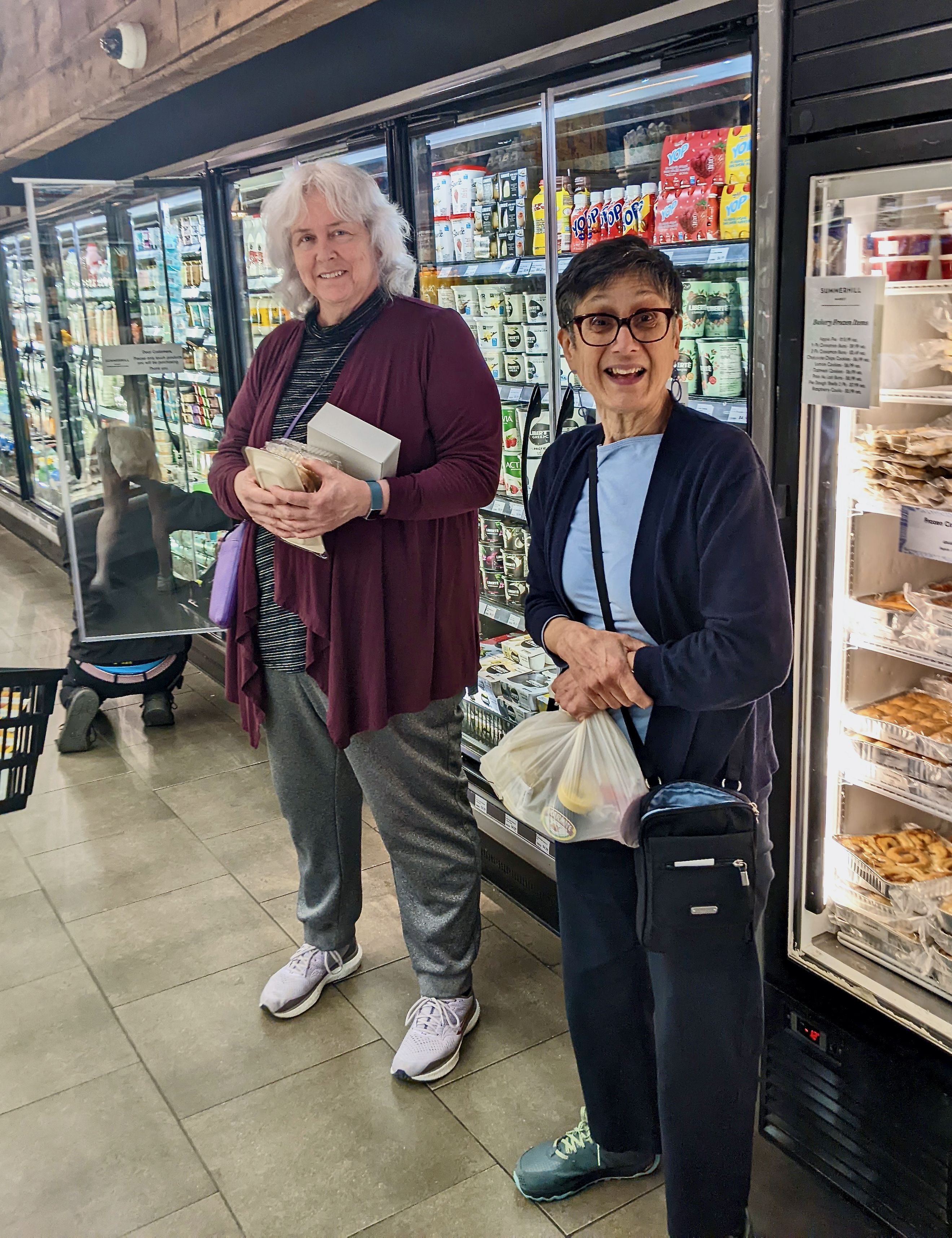 Two smiling women holding groceries standing in front of the frozen section in a grocery store.