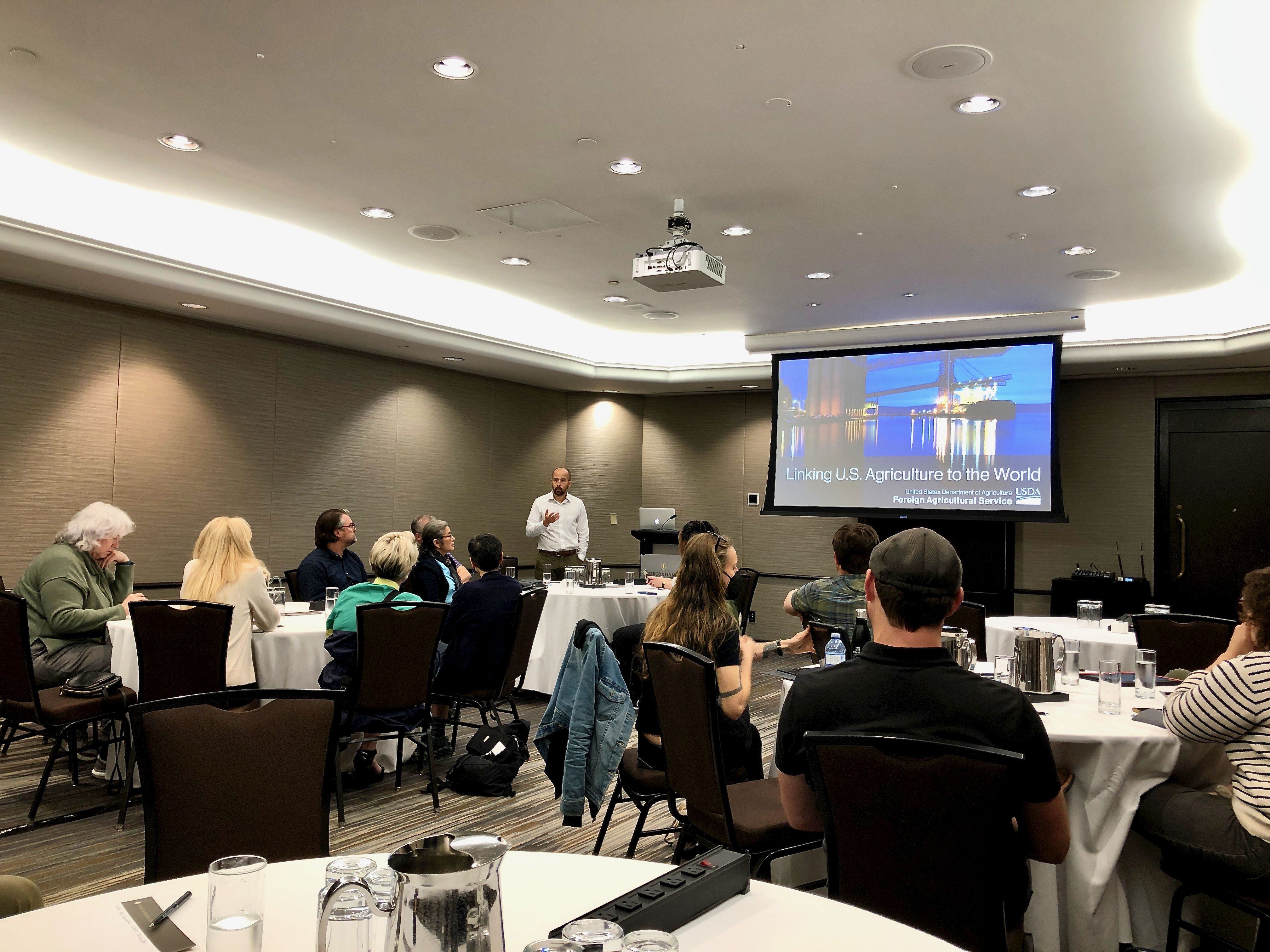 Man stands at the front of a hotel conference room with people sitting at round tables. A projector shows a slide on the screen that reads, "Linking U.S. Agriculture to the World"