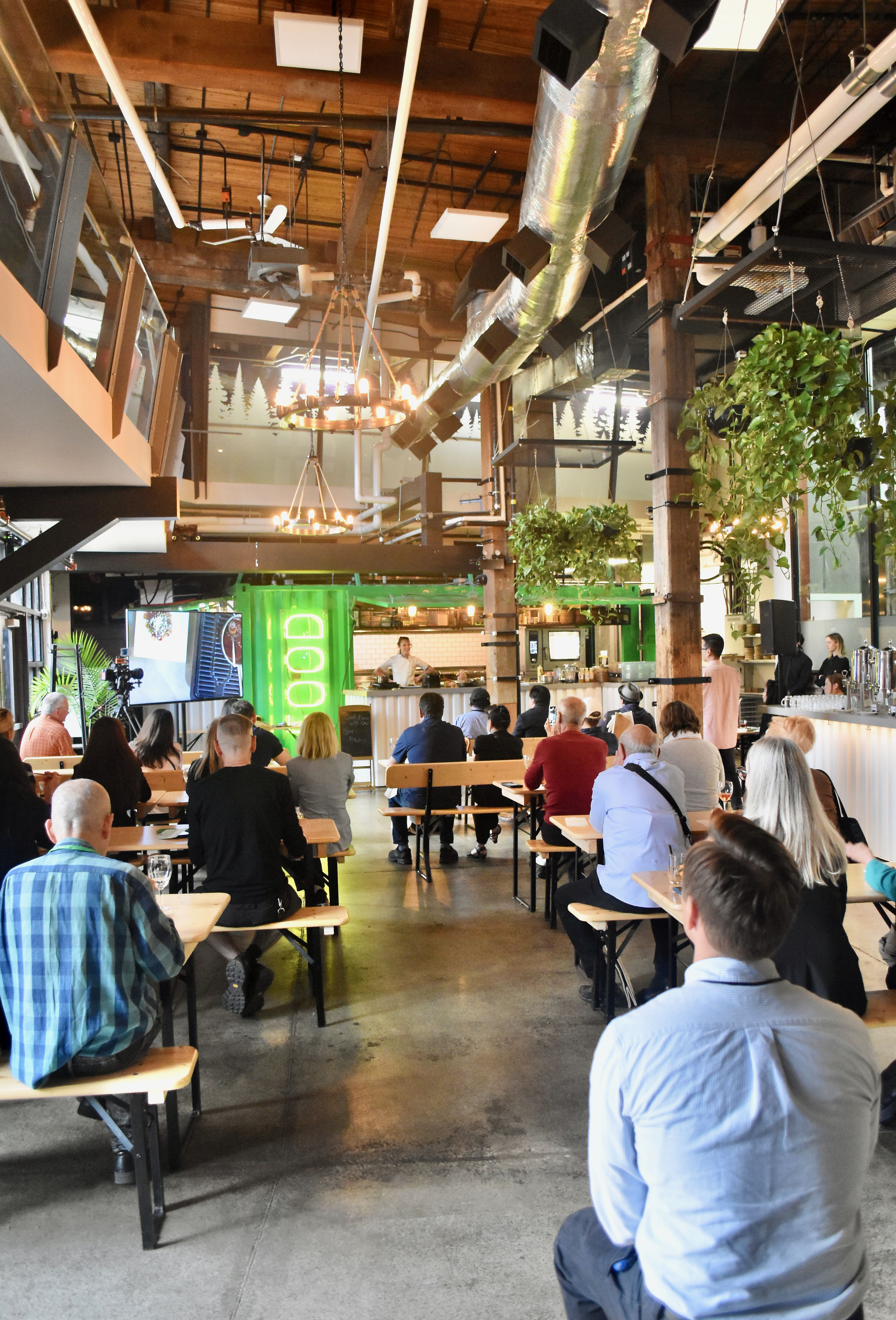 Looking down a hall with multiple tables and a kitchen at the back where people are watching a chef speak.
